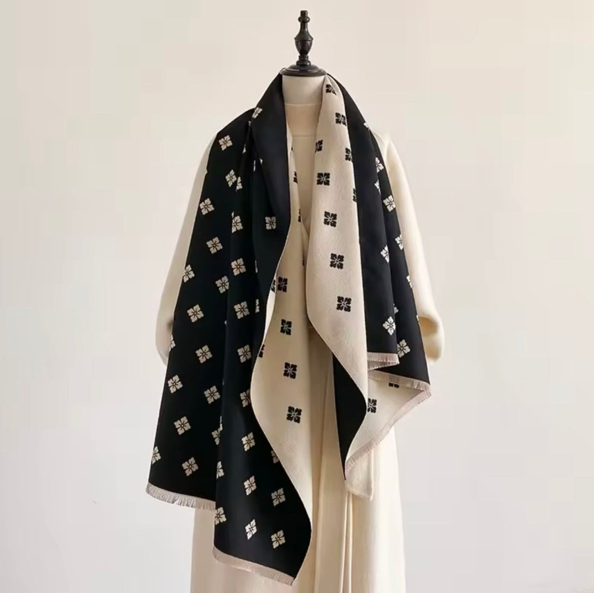 A black and cream silk scarf with a repeating geometric cross pattern, draped over a cream-colored dress form on a mannequin stand against a plain off-white background.