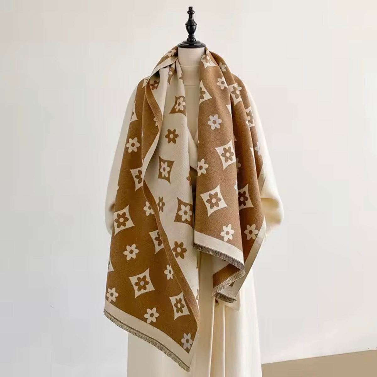 A brown and cream patterned shawl with white floral motifs is draped over a cream-colored dress form on a mannequin stand against a plain off-white background.