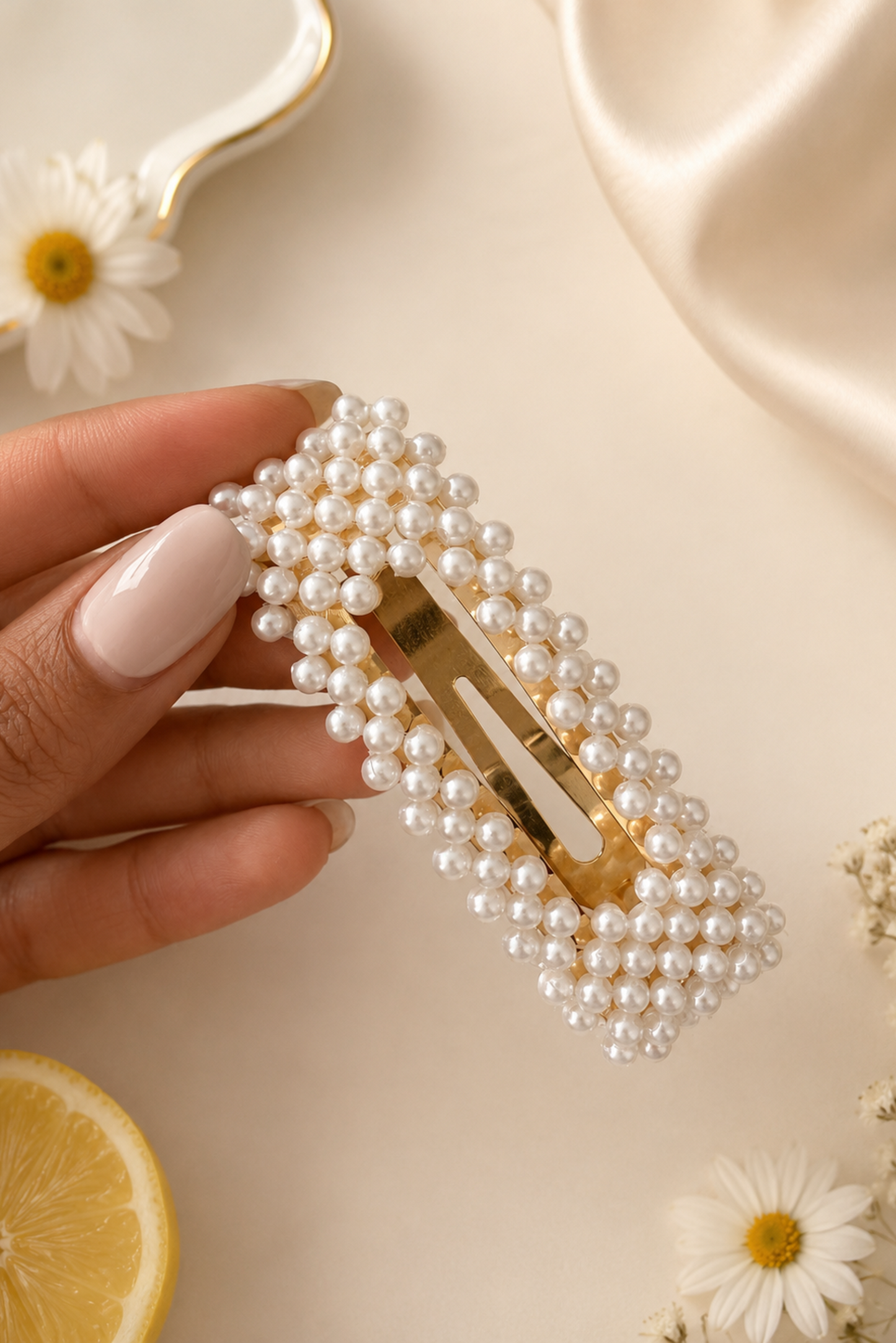 A hand with pale pink manicured nails holds a rectangular hair clip decorated with white faux pearls around a gold-tone metal center. The background is a warm cream color with decorative props including a lemon slice, daisies, and a satin fabric.