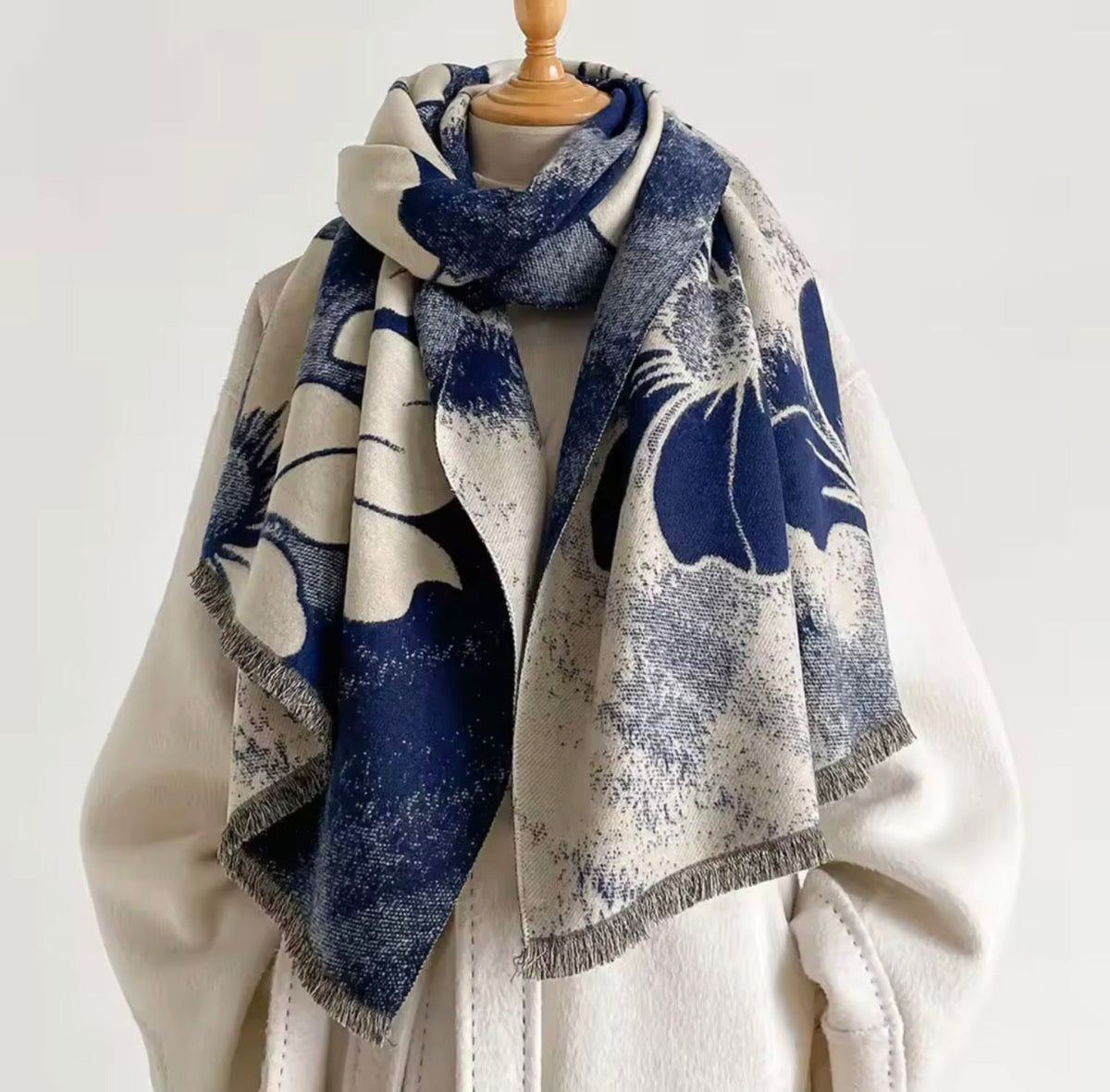 A large fringed scarf with a navy blue and cream abstract floral print, draped on a cream-colored coat displayed on a mannequin torso against a plain light background.