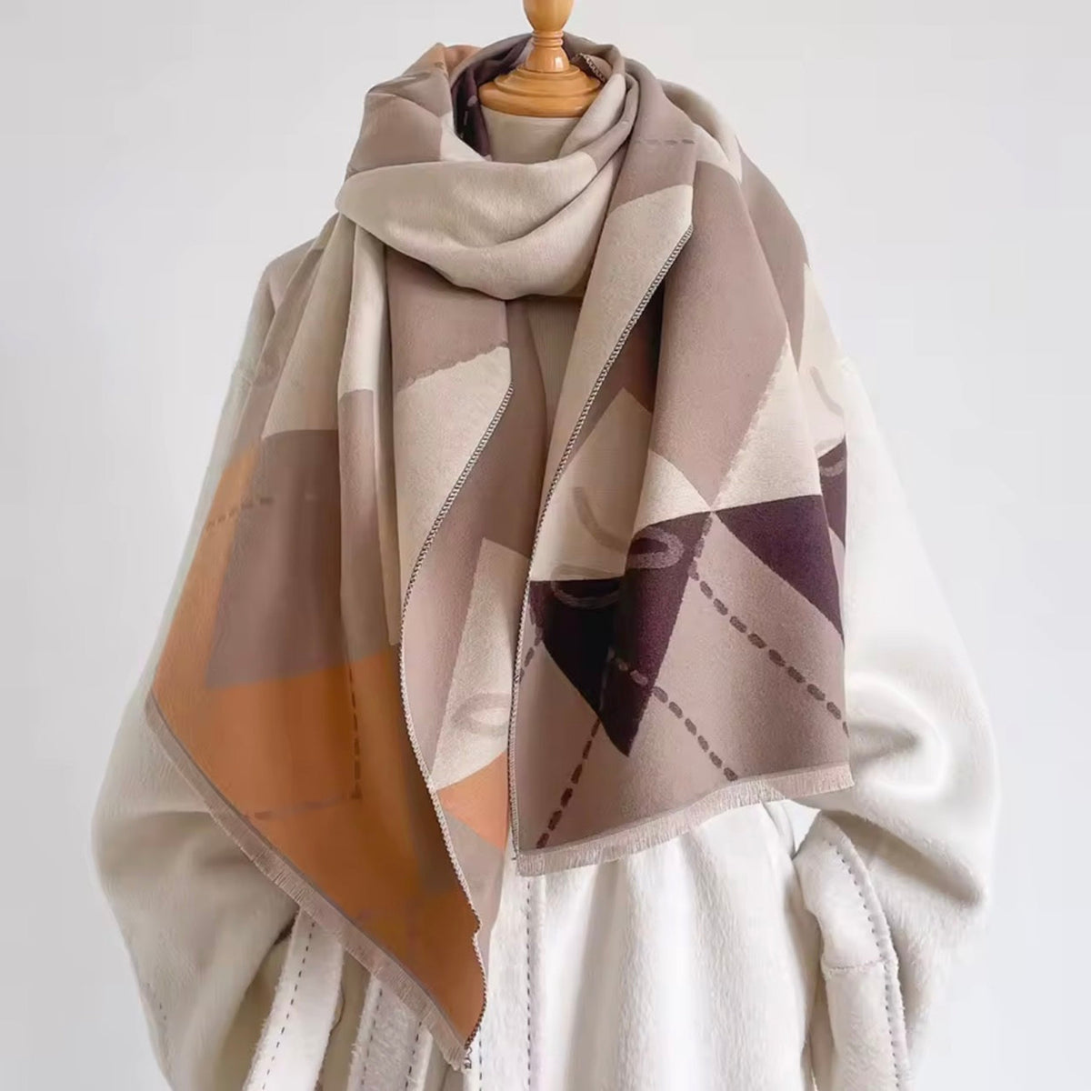 A large geometric patterned scarf draped over a cream-colored coat on a wooden mannequin torso, against a plain light gray background.