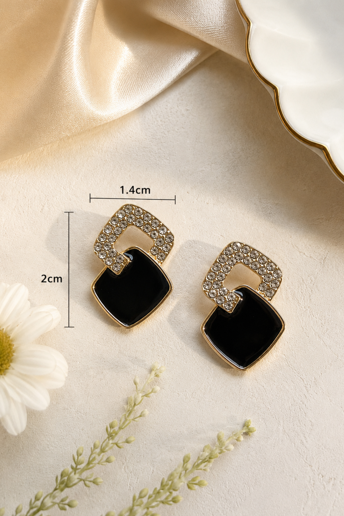 A pair of gold-tone statement earrings with a black square enamel drop and a curved top section paved with small clear rhinestones, shown on a beige fabric background with decorative dried flowers and a white dish. Measurement annotations indicate 1.