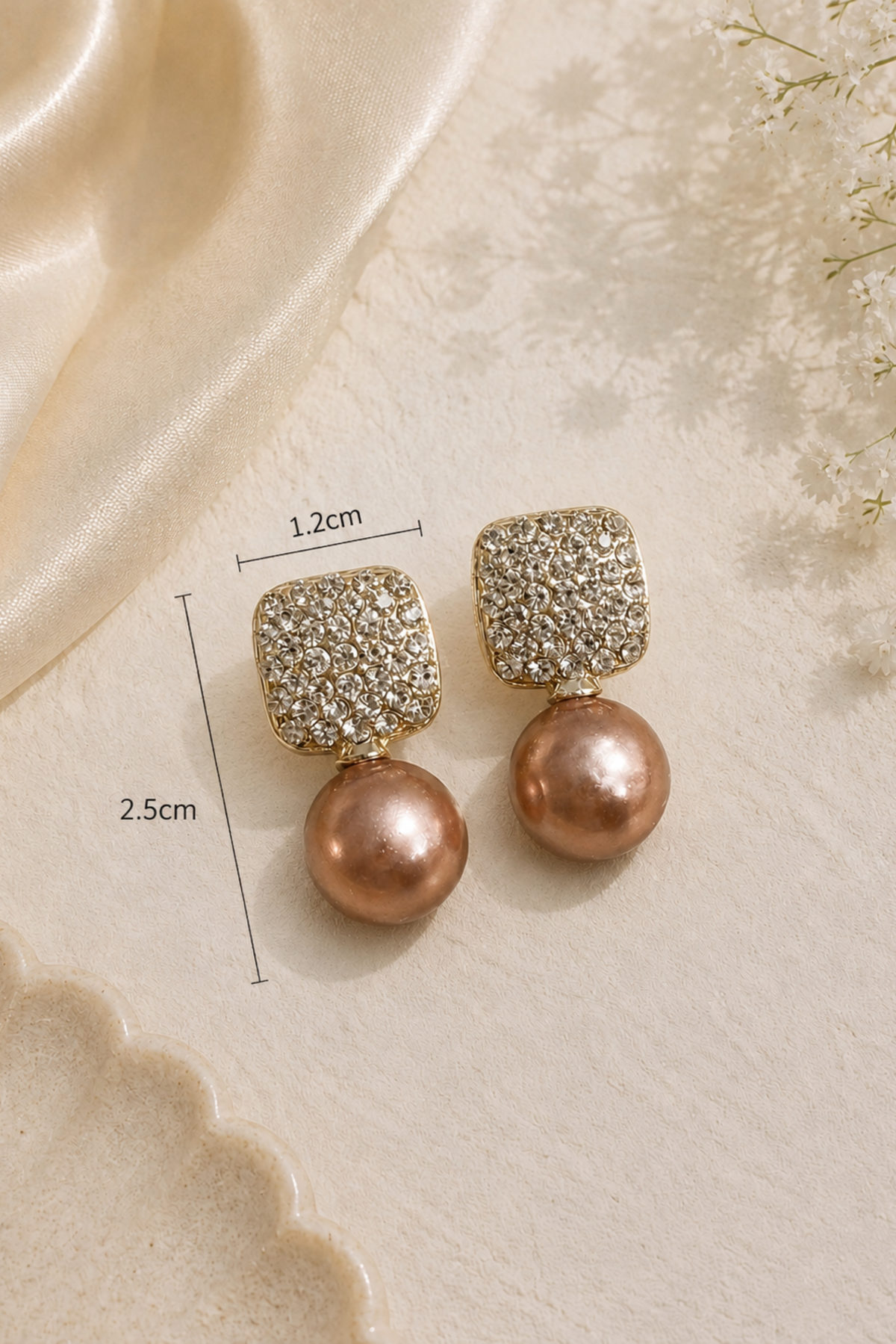 A pair of gold-toned drop earrings with square crystal-encrusted tops and round rose-gold pearl drops, shown on a beige textured surface with satin fabric and small white flowers. Measurement lines indicate 1.