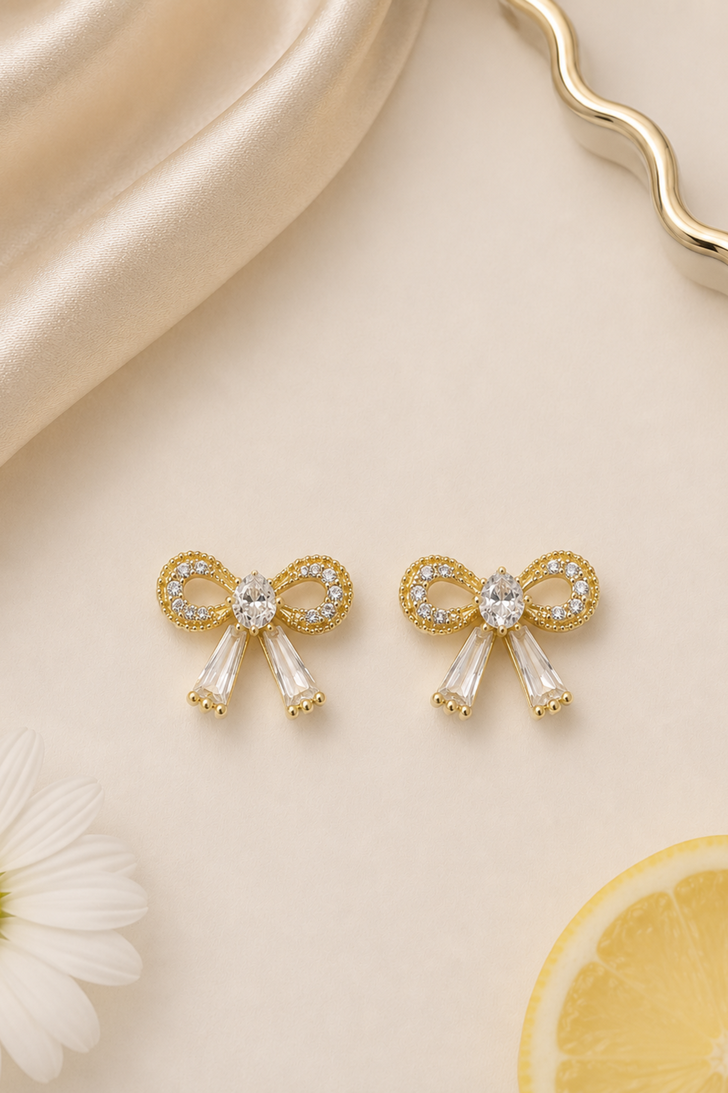 A pair of gold bow-shaped stud earrings with clear crystal accents, displayed on a cream-colored surface with a daisy flower, a lemon slice, and draped satin fabric.