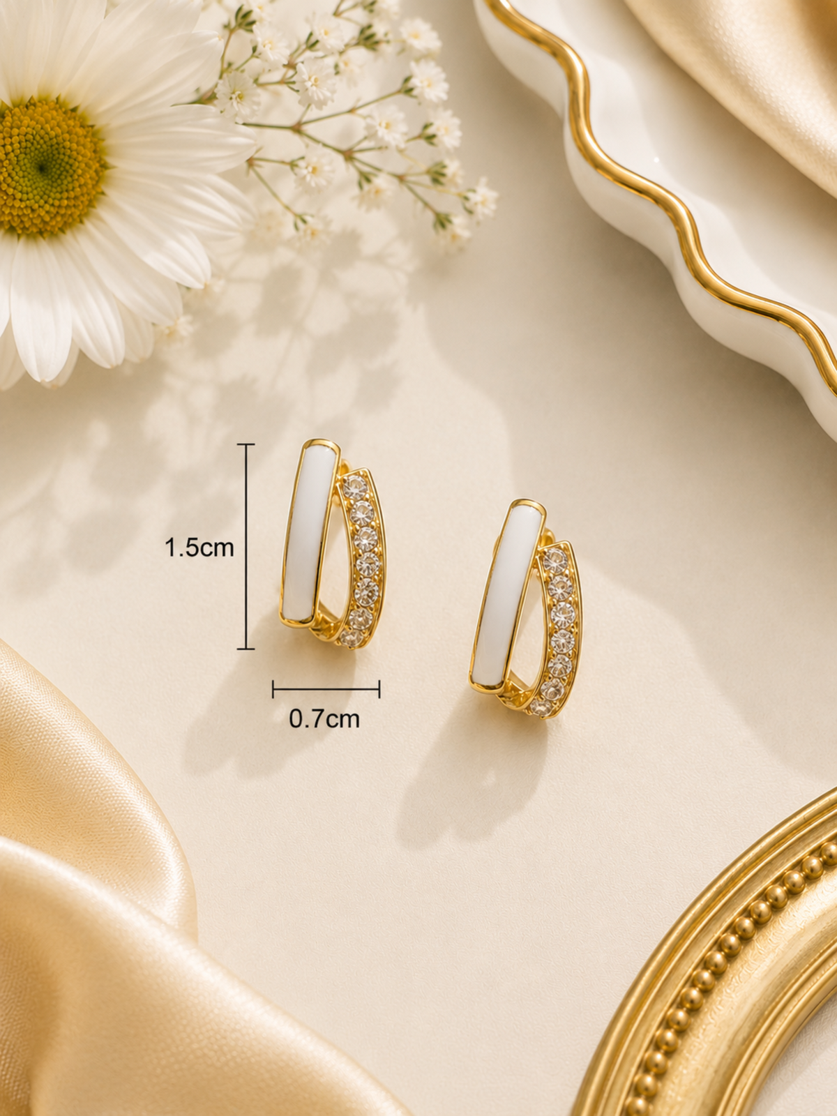 A pair of gold hoop-style earrings with white inlay and small clear stones, shown on a beige surface with decorative flowers, satin fabric, and a mirror edge. Measurement labels indicating 1.