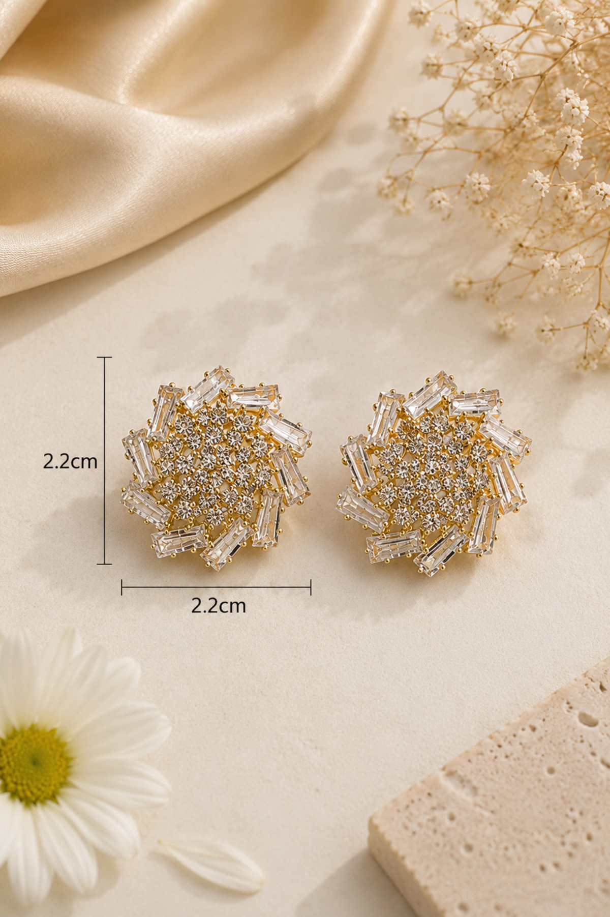 A pair of round, flower-shaped stud earrings with layered clear crystal petals and gold-toned centers, shown on a light beige surface with decorative satin, dried flowers, a daisy, and a stone slice. Measurement lines indicate the earrings are 2.
