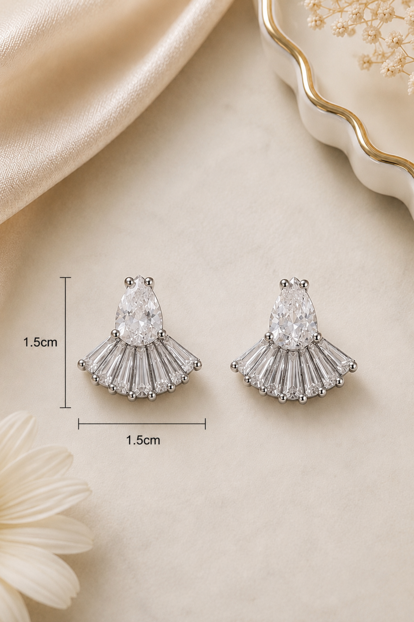 A pair of silver-tone fan-shaped stud earrings with clear marquise and baguette stones, shown on a cream surface with decorative satin and a dish. Black measurement lines and text indicate 1.