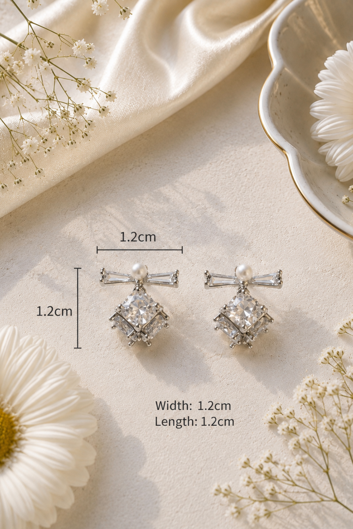 A pair of silver-toned bow and flower drop earrings with clear rhinestones and pearl-like center beads, shown on a cream surface with decorative daisies, baby's breath, and draped satin. Measurement lines and text indicate the earrings are approximately 1.