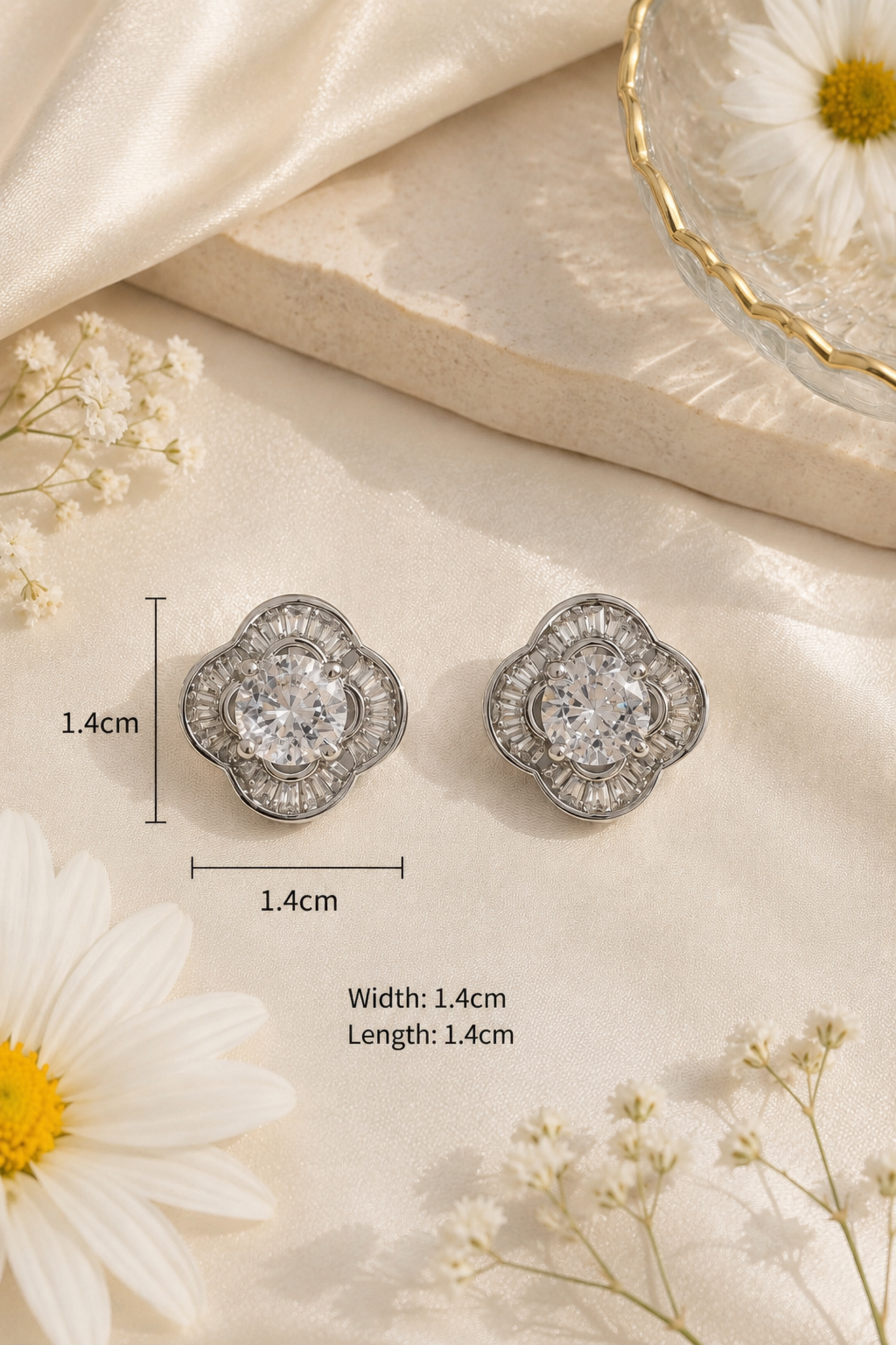 A pair of silver-toned floral stud earrings with clear central stones, shown on a cream satin surface with daisy flowers and small white blossoms. Measurement lines and text indicate the earrings are approximately 1.