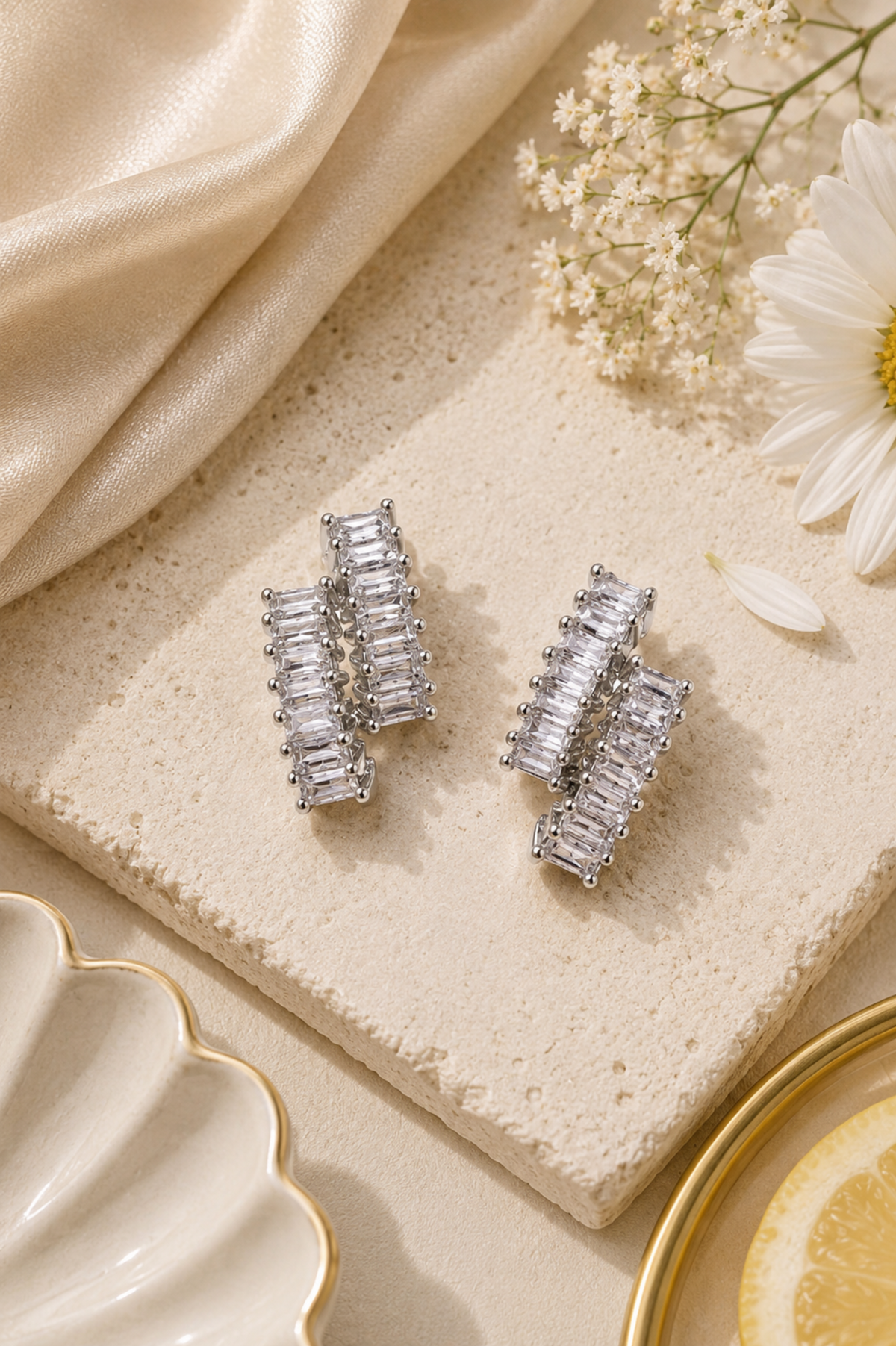 A pair of silver-toned rectangular baguette stud earrings displayed on a textured beige stone slab with cream satin fabric, white flowers, and gold-toned dish edges in the background.