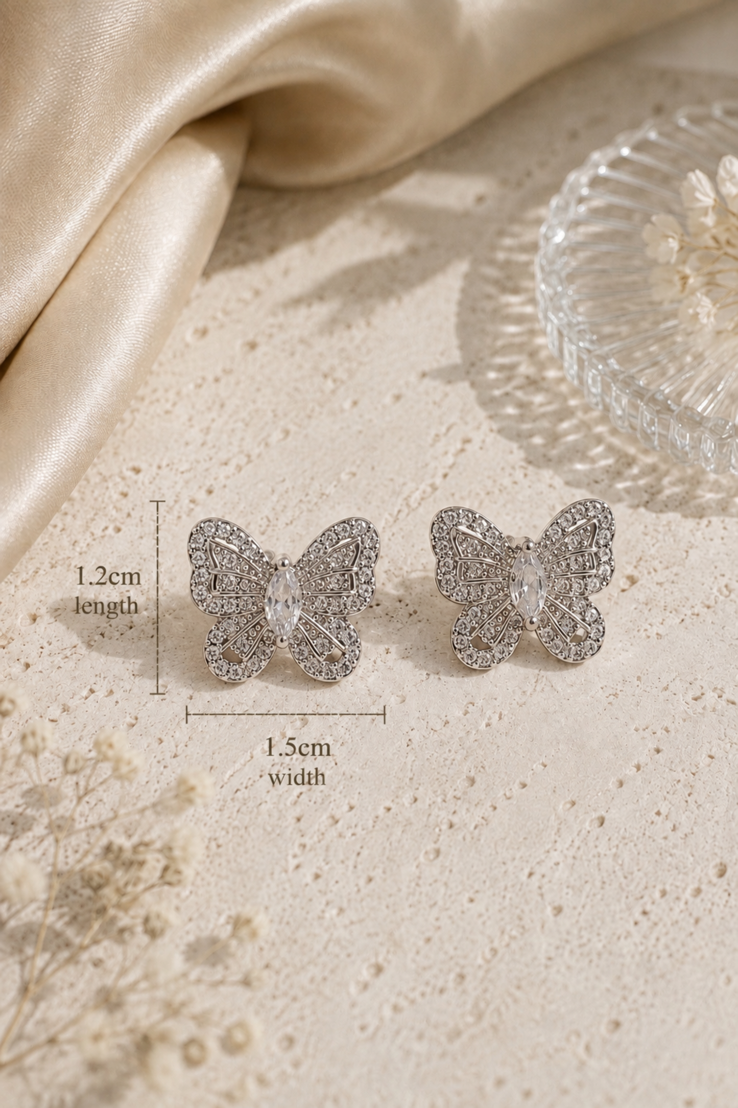 A pair of silver butterfly stud earrings with intricate crystal detailing, displayed on a textured beige surface with dimension lines indicating 1.2cm length and 1.