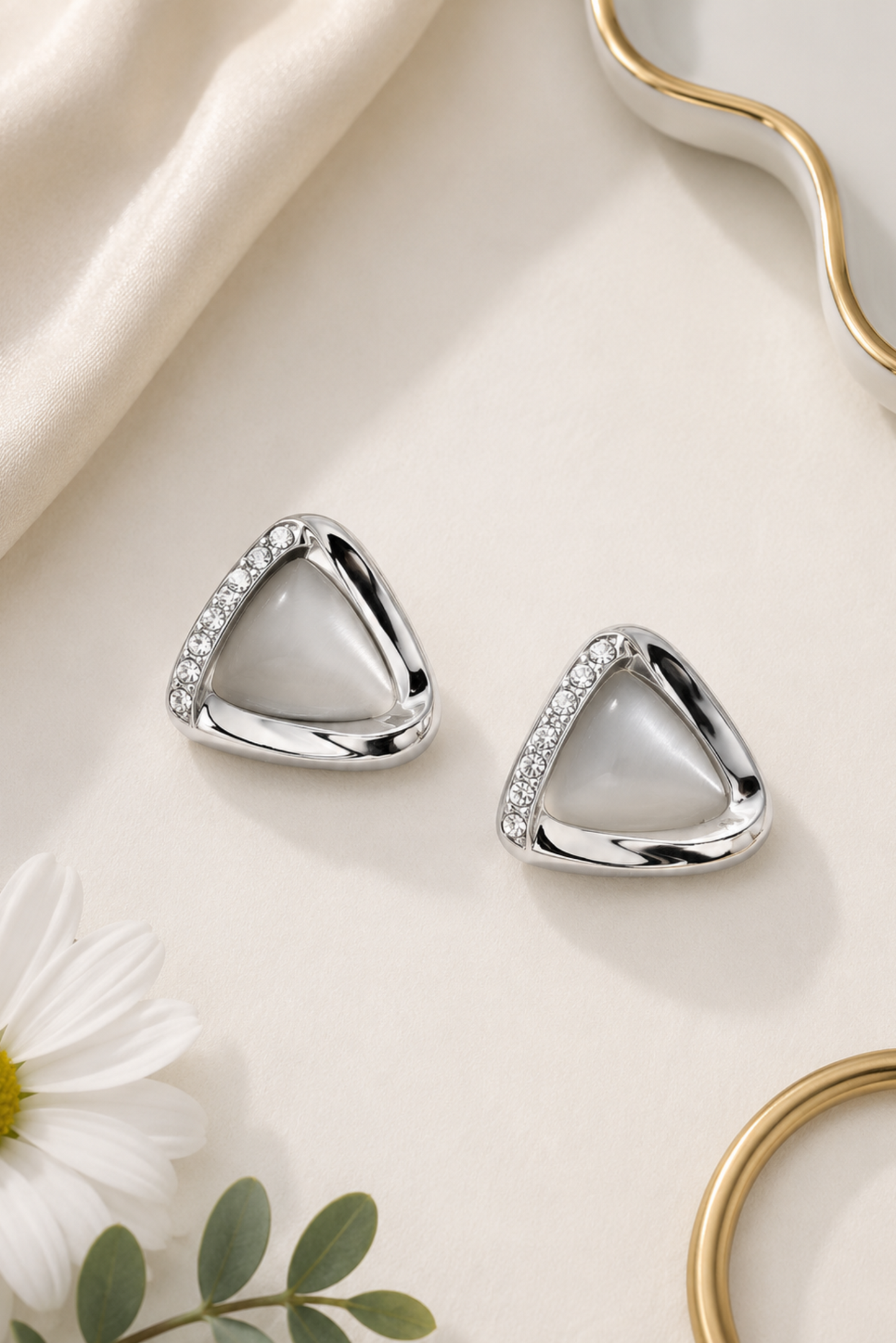 A pair of silver triangle stud earrings with a smooth white center and a row of small clear stones along one side, displayed on a cream surface with decorative fabric, a daisy flower, and gold-toned props.