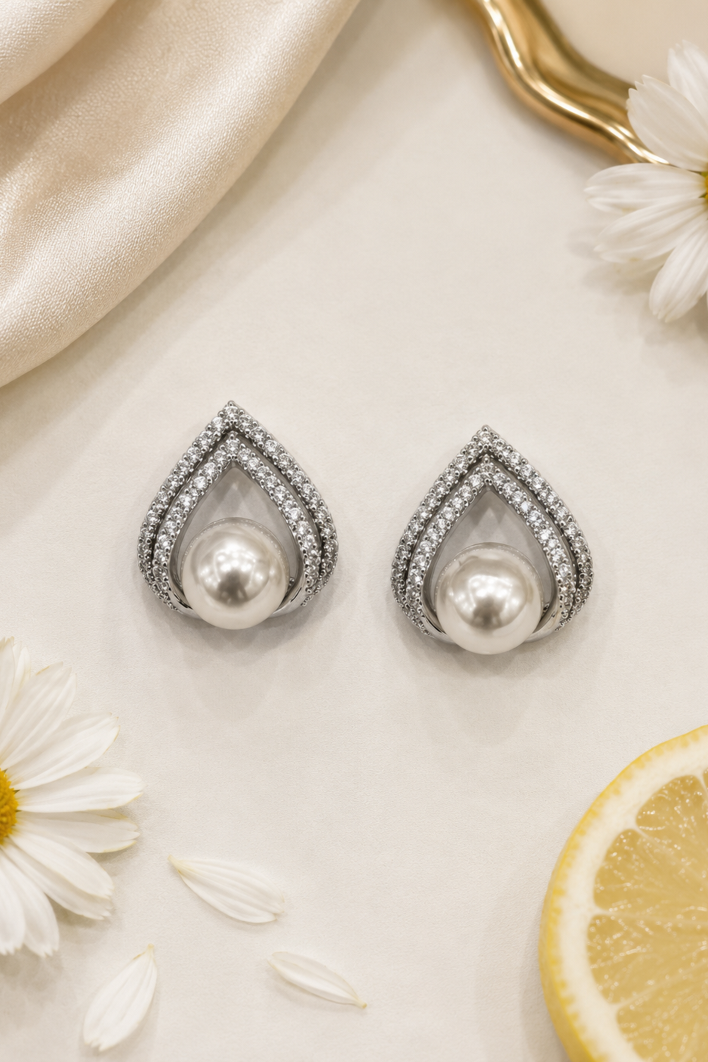 A pair of teardrop-shaped pearl earrings with silver pavé borders, styled on a cream surface with white daisies, a lemon slice, and a gold spoon.