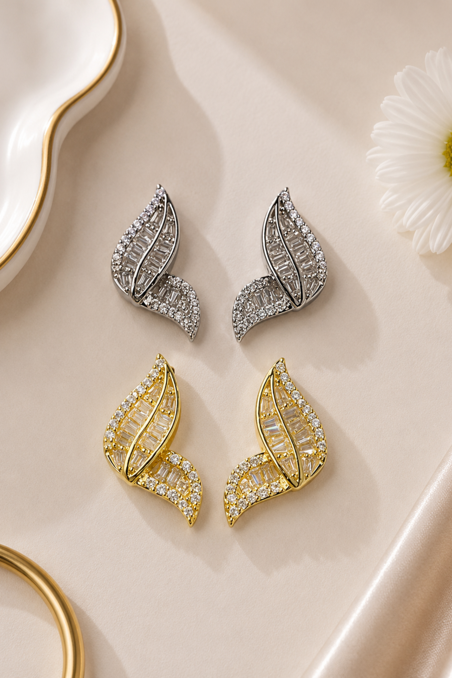 Four leaf-shaped stud earrings arranged on a warm beige surface, with two silver-toned earrings and two gold-toned earrings, each set with clear stones. A white flower and partial decorative dishes are visible as styling props.