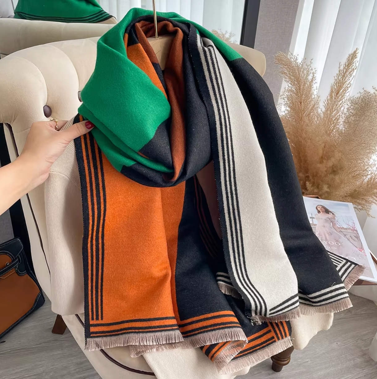 A green and orange striped wrap scarf shawl draped over a chair with a person's hand adjusting the green striped end.
