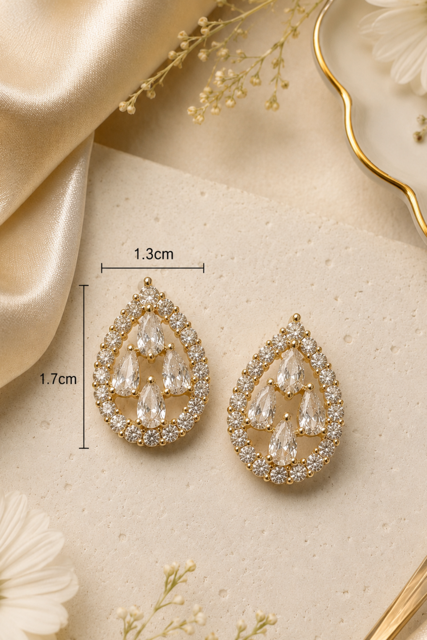 Teardrop-shaped gold statement earrings with clear crystal drops, displayed on a textured beige surface with decorative dried flowers and gold satin. Measurement overlays indicate 1.