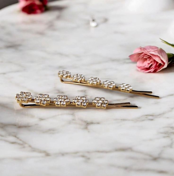 Two gold hair pins with crystal embellishments on a marble surface with pink roses.