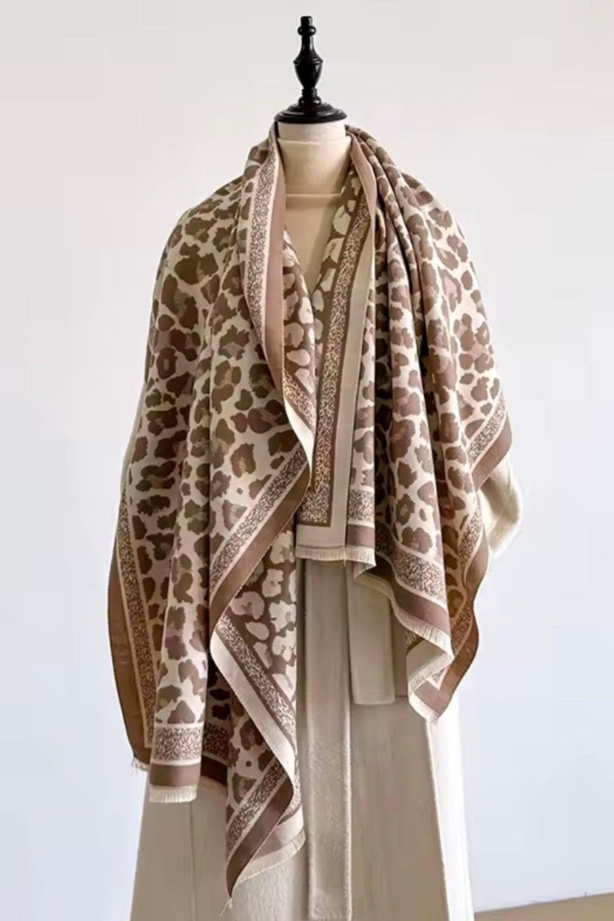 A beige and brown animal-print shawl with ornate border patterns draped over a cream-colored dress form against a plain white wall.