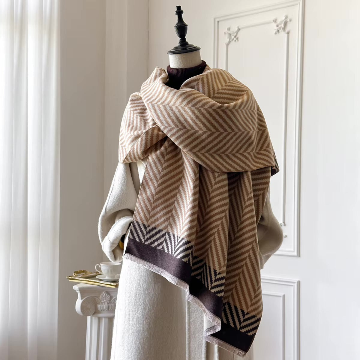 A beige and brown herringbone patterned scarf with a dark brown geometric border, draped on a cream-colored coat dress form in a bright, elegant room.