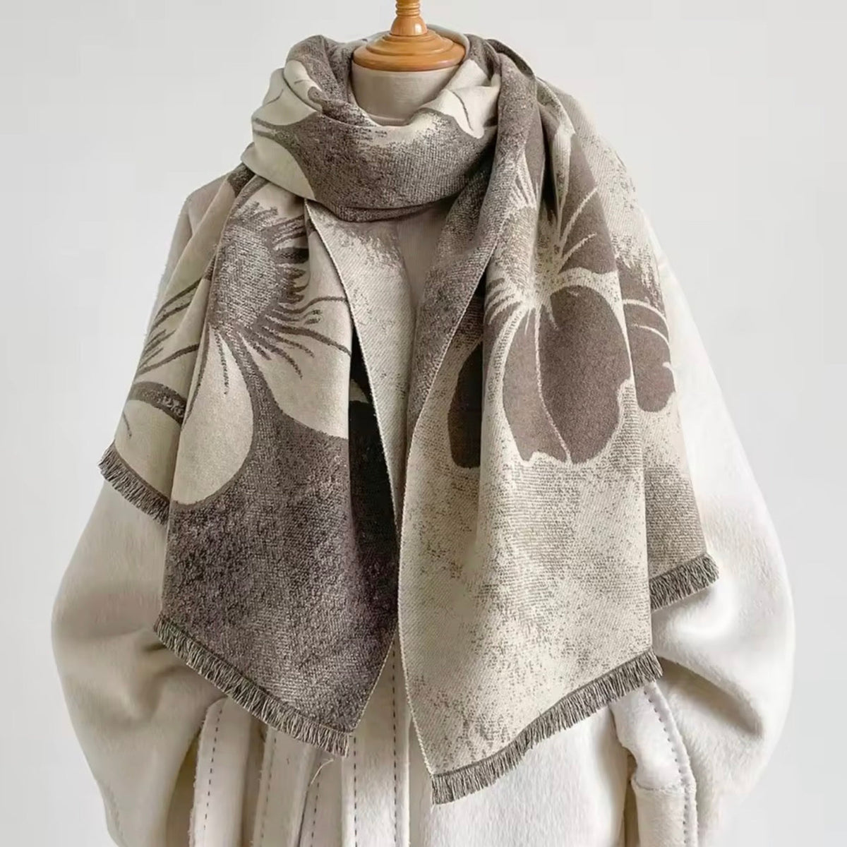 A beige and brown patterned scarf with fringed edges draped over a cream-colored coat on a mannequin torso against a light gray background.