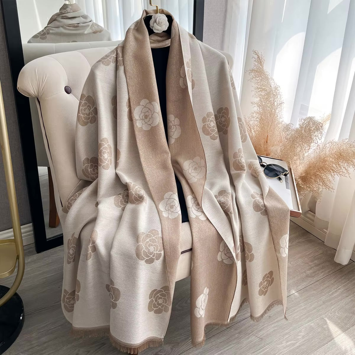 A beige and cream floral shawl with fringed edges draped over a cream tufted armchair in a bright, modern room.