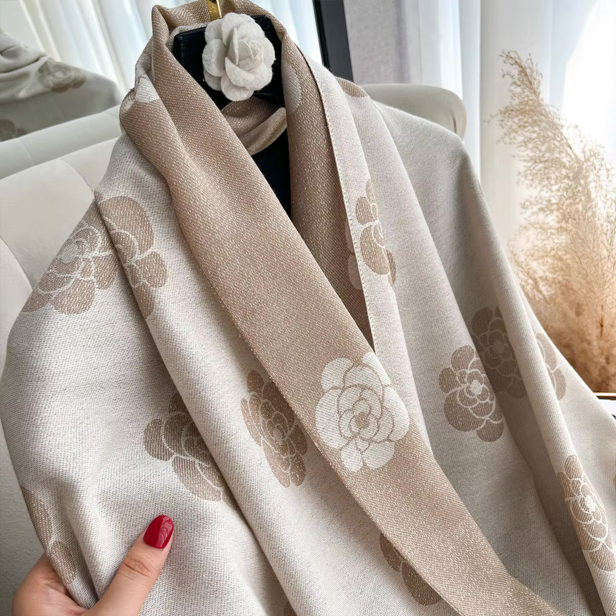 A beige and off-white scarf with a tonal floral camellia pattern draped over a hanger, held by a hand with red nail polish, in a softly lit interior setting.