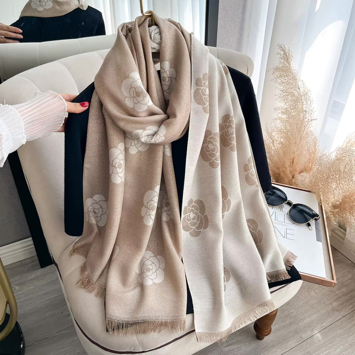 A beige and off-white scarf with large floral patterns draped over a cream-colored armchair in a bright, modern room. A hand in a white ribbed sleeve holds the left side of the scarf, and sunglasses and a tray are placed on the chair beside it.