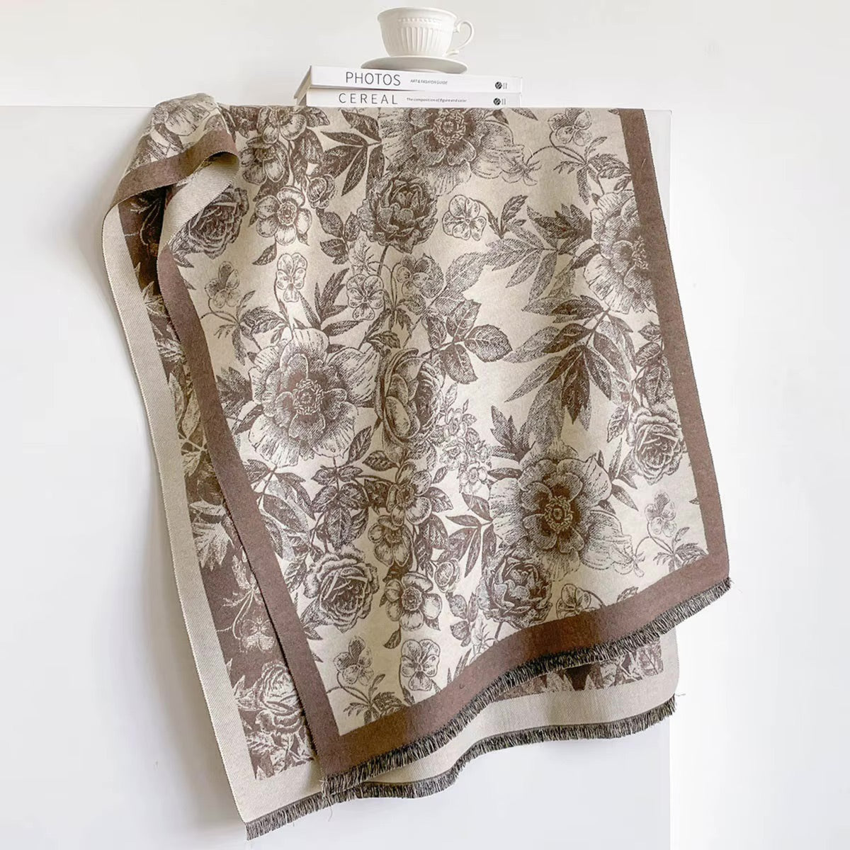 A beige scarf with a brown floral rose pattern and fringed edges, draped over a white surface with a white book and cup in the background.