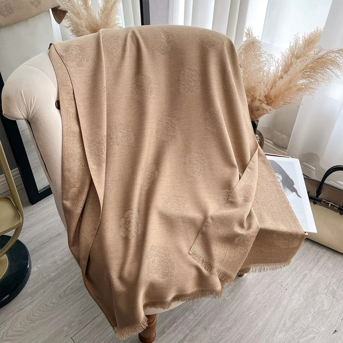 A beige shawl with a subtle tonal damask pattern draped over a cream-colored armchair in a bright, modern room.