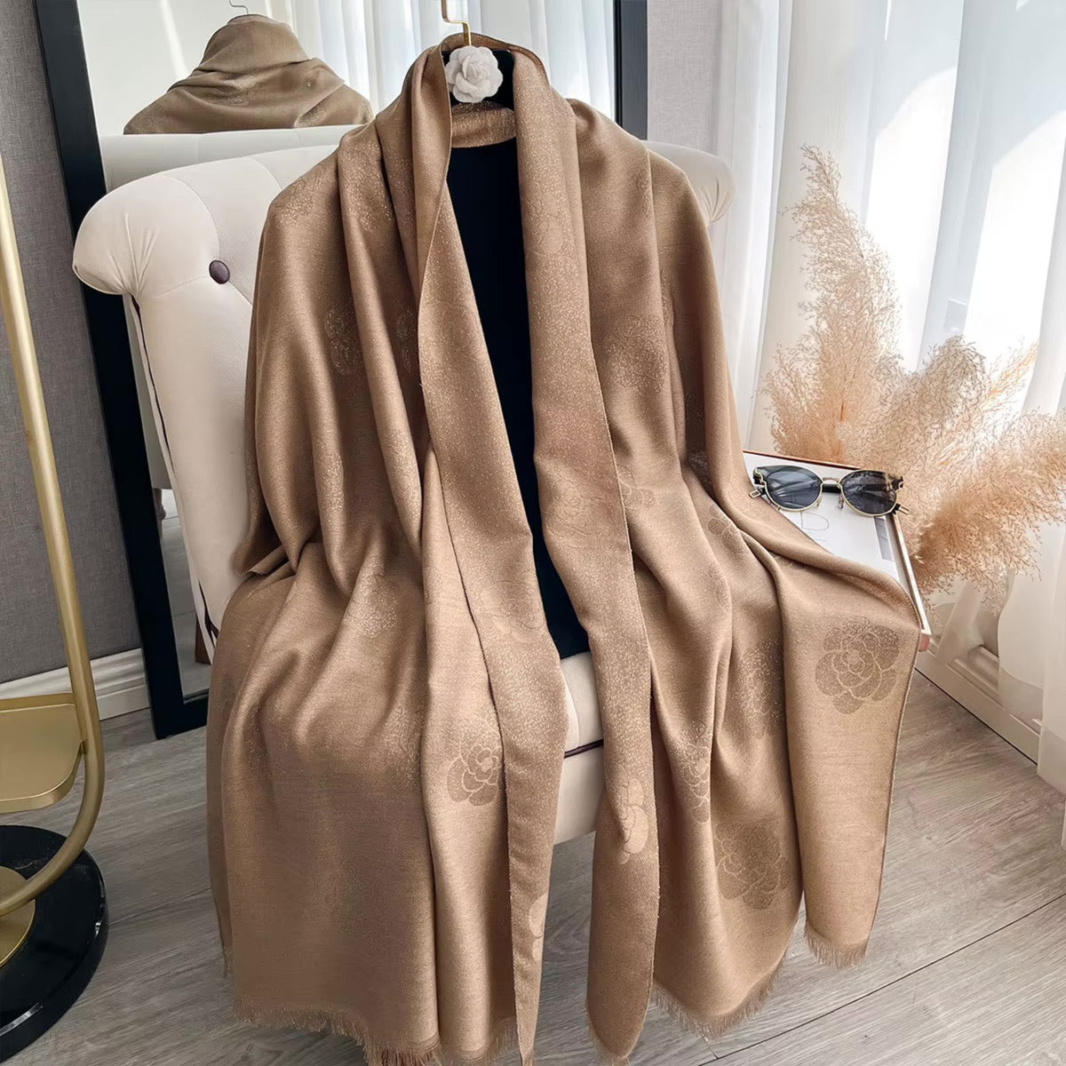 A beige shawl with subtle floral jacquard patterns draped over a black top on a hanger, styled in a bright, modern room next to a cream tufted chair and pampas grass.