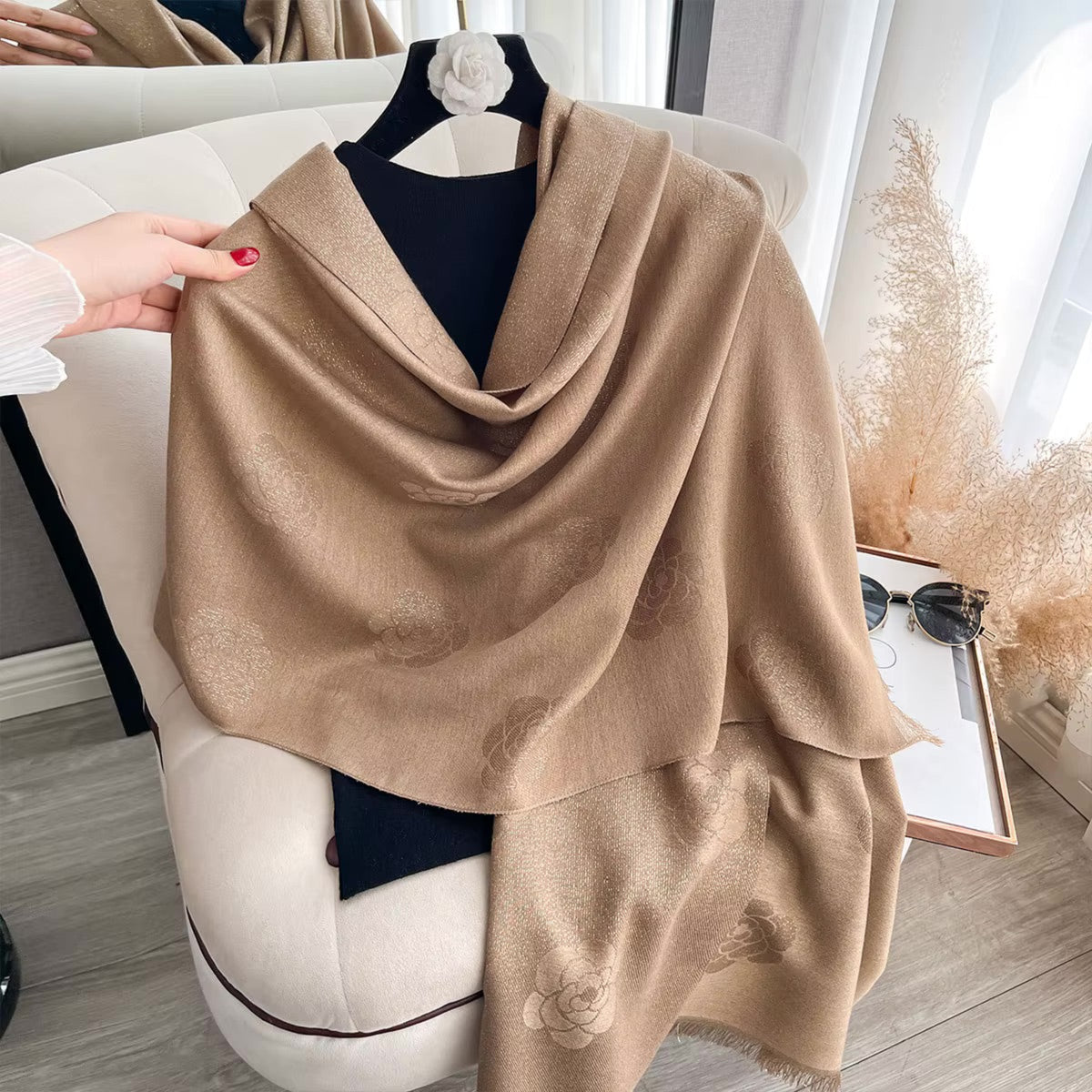 A beige shawl with subtle tonal floral jacquard patterns draped over a black top on a hanger, styled on a cream-colored armchair with a hand holding its edge.
