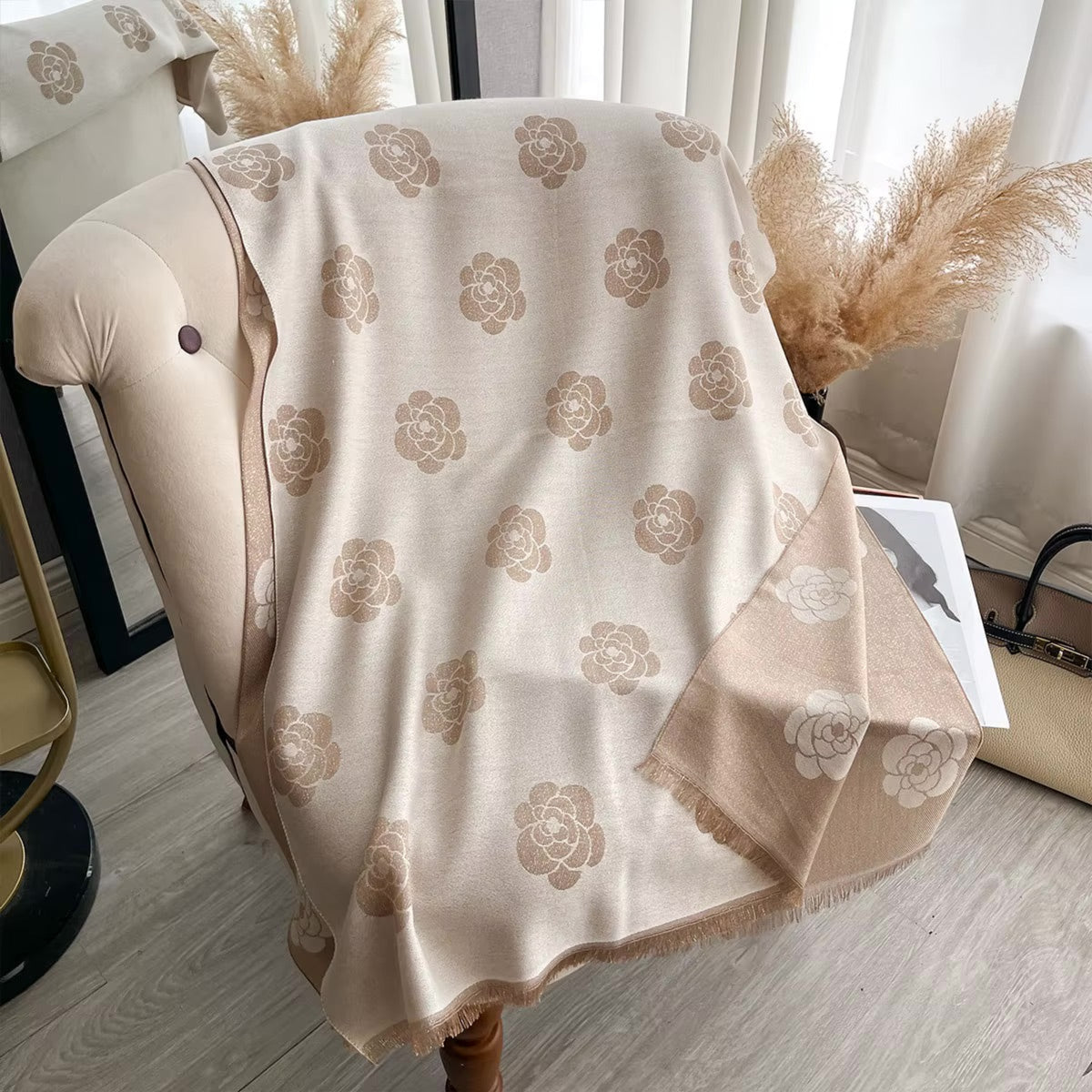 A beige throw blanket with a repeating tan floral pattern draped over a tufted armchair in a styled interior setting.
