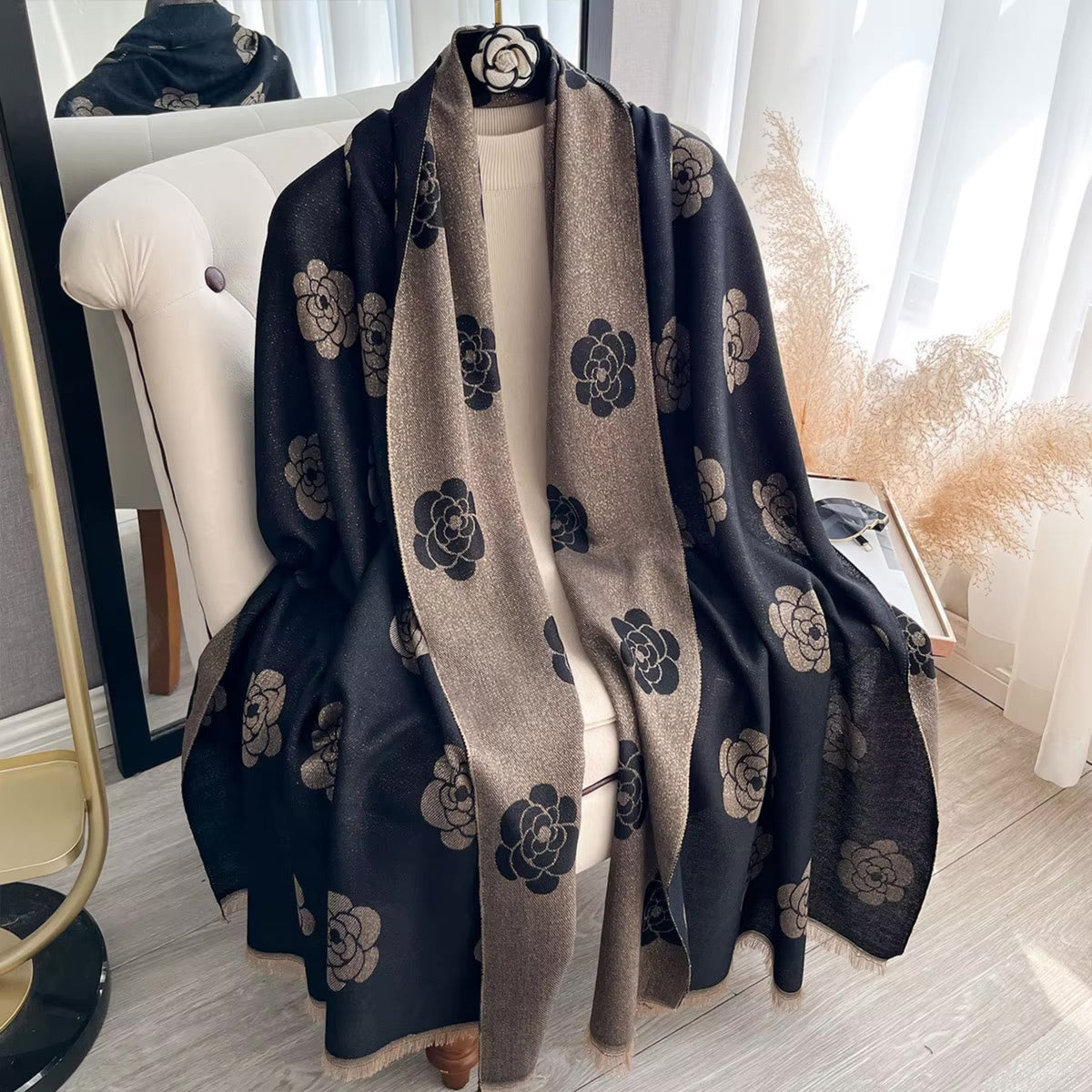 A black and beige floral shawl draped over a cream-colored chair in a bright, modern room with light wood floors and pampas grass decor.