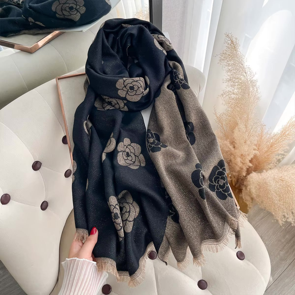 A black and taupe floral scarf with fringed edges draped over a cream tufted armchair, with a hand visible at the bottom left holding it in place.