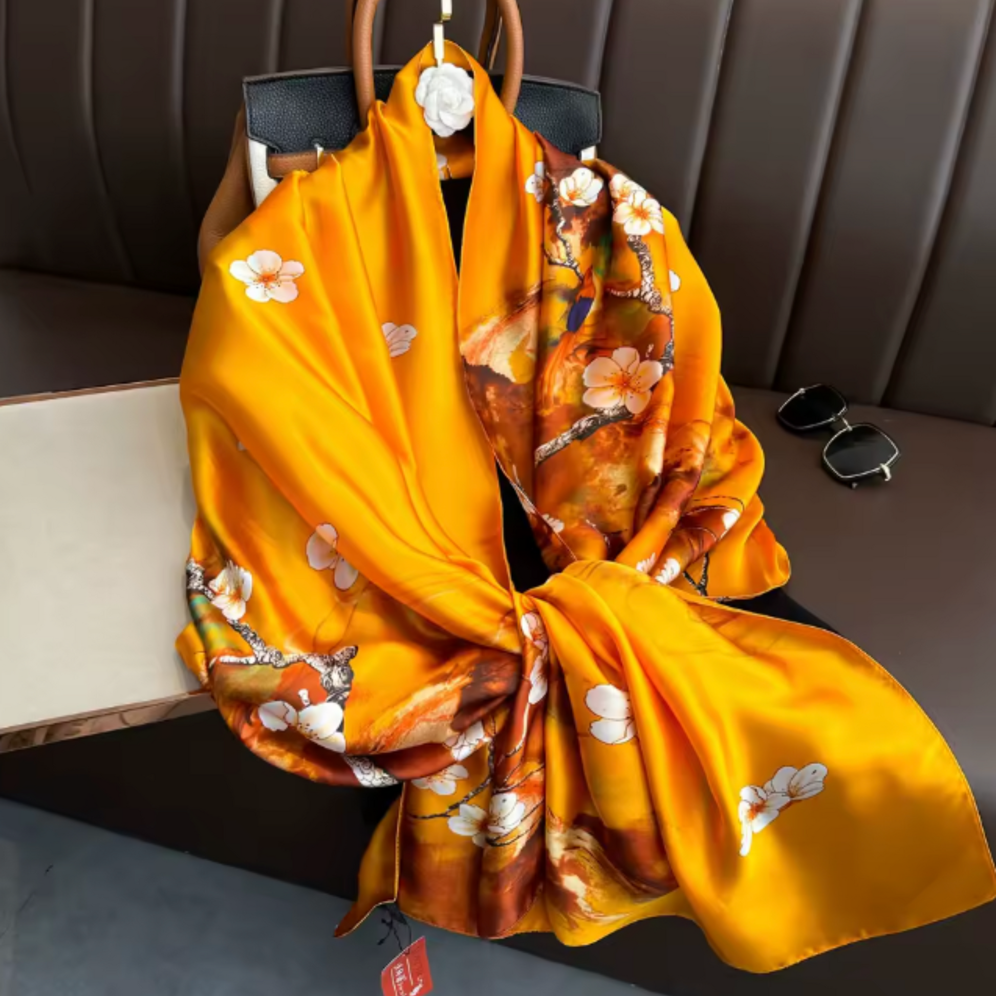 A bright yellow-orange silk scarf with a white floral print draped over a brown leather handbag, with sunglasses resting on the brown leather seat next to it.