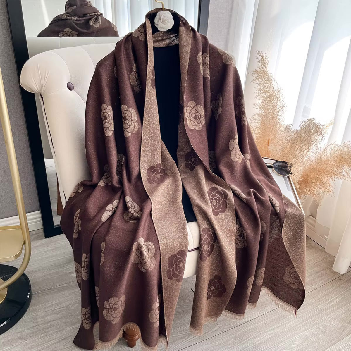 A brown and beige patterned shawl with large floral motifs draped over a white tufted chair in a bright indoor setting.