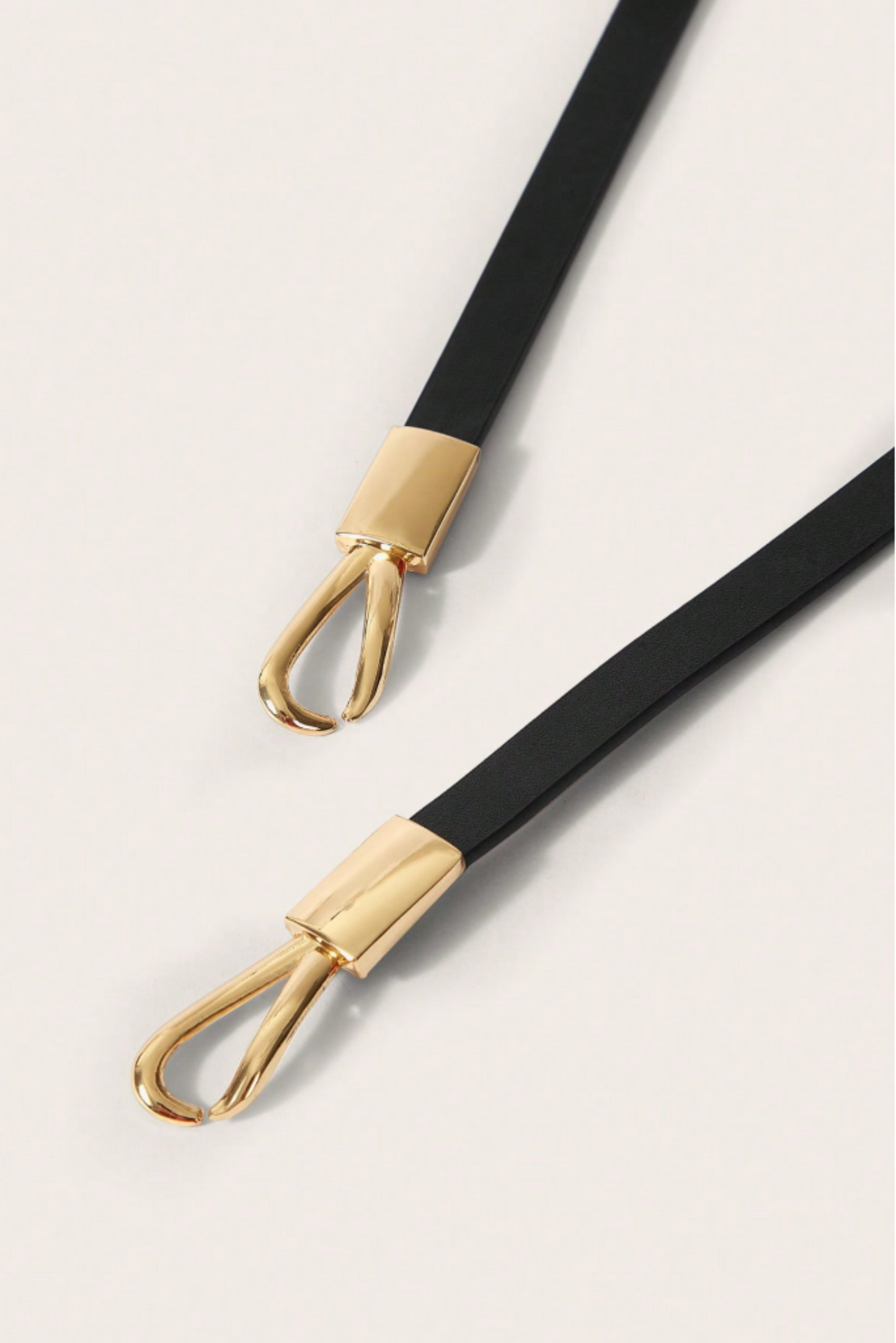 A close-up of a black strap with polished gold-tone metal hardware, shown diagonally on a light off-white background.
