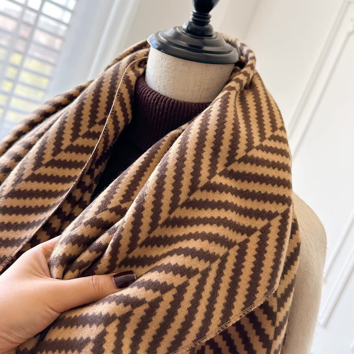 A close-up of a brown and beige chevron-patterned scarf draped on a mannequin torso, with a hand holding the fabric to show its texture.
