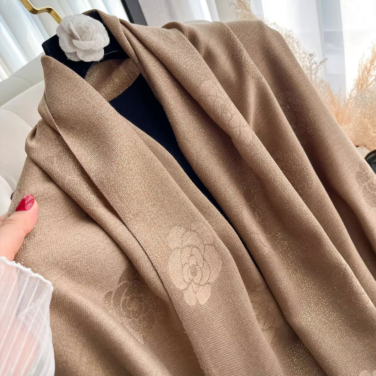 A close-up of a draped taupe-brown shawl with a subtle tonal floral jacquard pattern, styled on a black hanger with a white fabric flower accent.