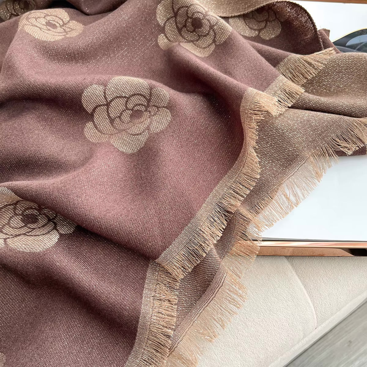 A close-up of a dusty mauve-brown woven throw blanket with beige floral motifs and fringed edges, draped over a light-colored surface.