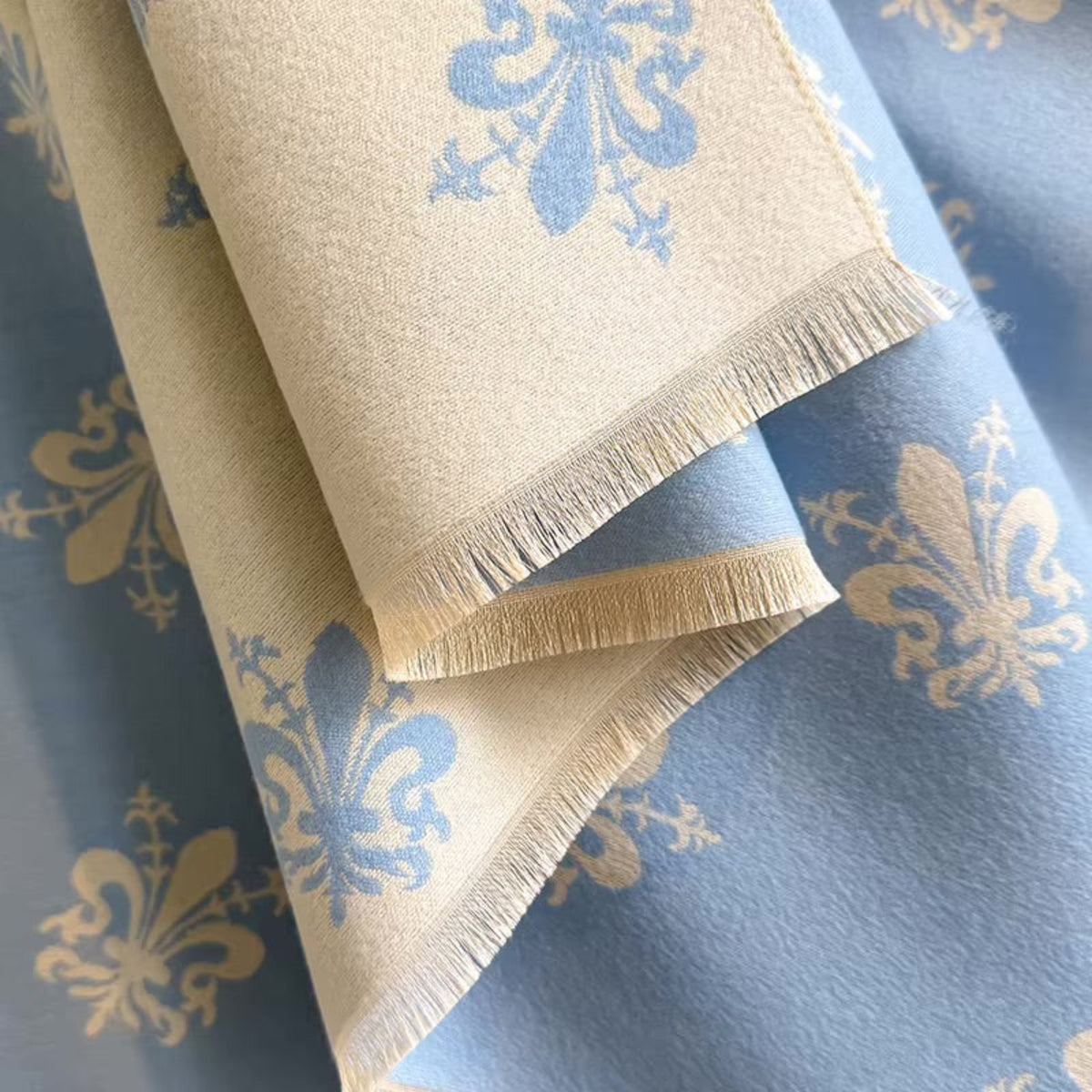 A close-up of a folded woven textile with a cream base and light blue floral pattern, shown over a matching blue fabric with gold floral motifs.