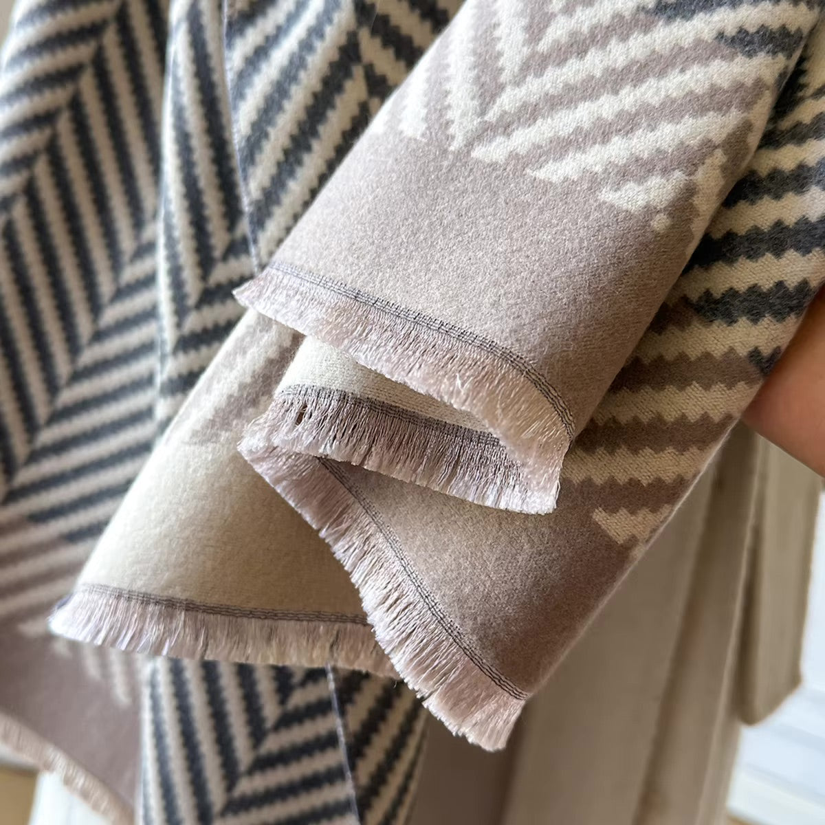 A close-up of a folded woven throw blanket with fringed edges, showing a soft geometric pattern in taupe, beige, and dark charcoal gray. A hand is holding the blanket, and another matching blanket is draped in the background.