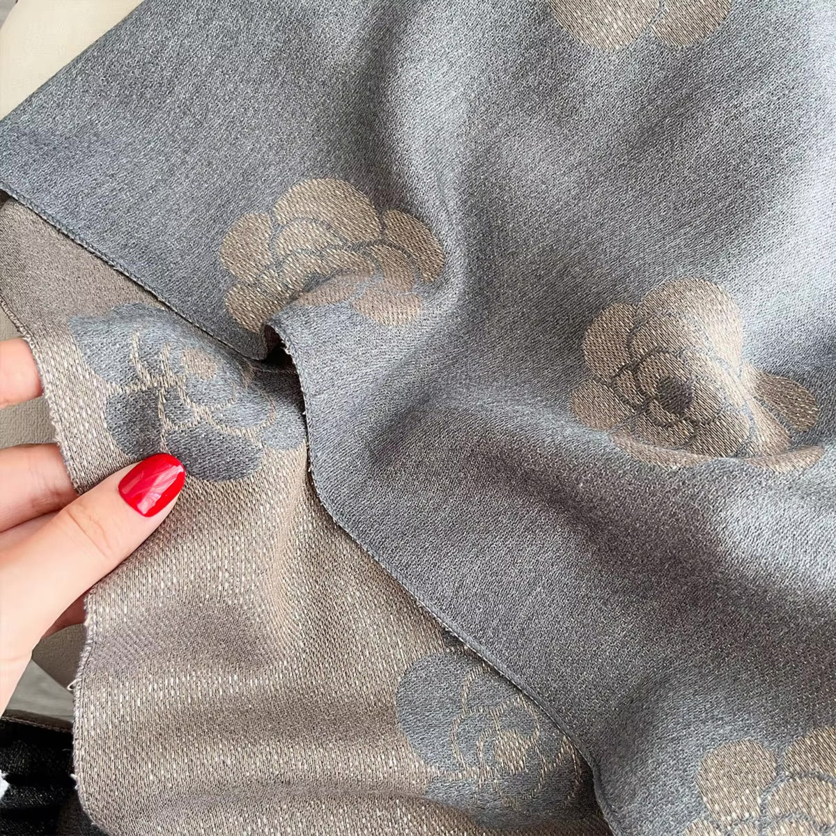 A close-up of a gray fabric with a woven floral pattern in beige and gray, held by a hand with red painted fingernails.