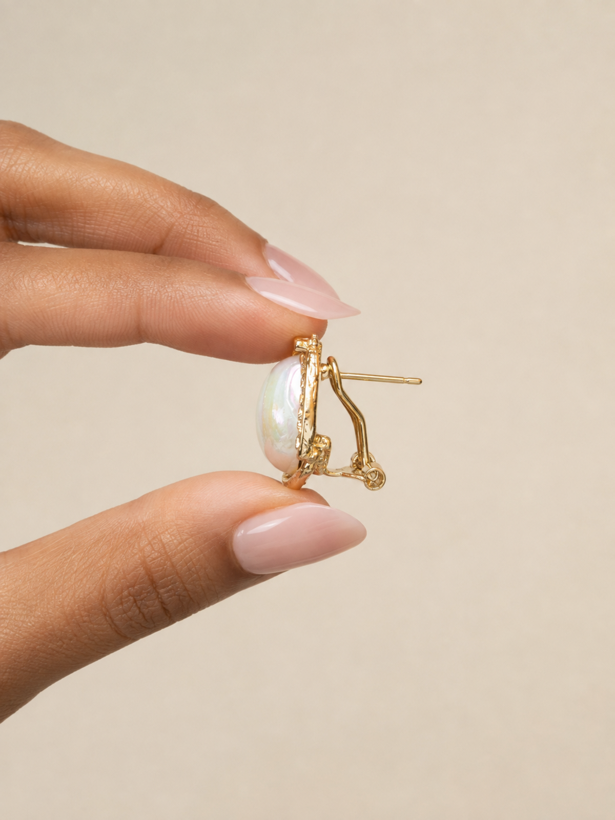 A close-up of a hand with pale pink manicured nails holding a gold hoop earring with an oval iridescent white pearl against a solid beige background.