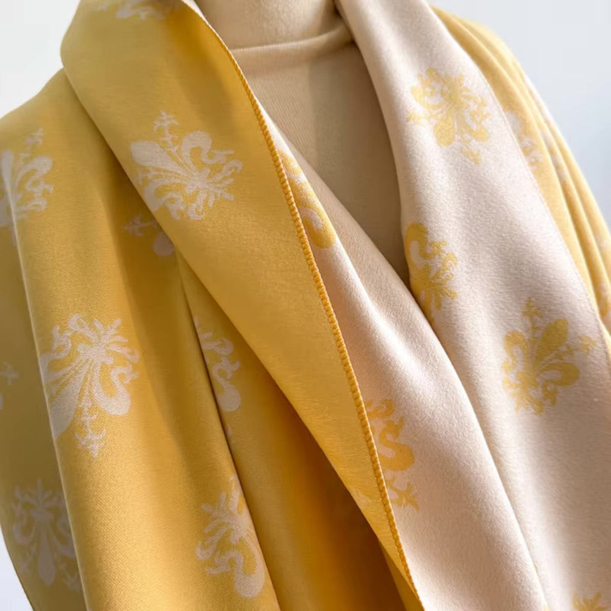 A close-up of a lightweight scarf draped on a mannequin, showing two main colors: a golden yellow side with white damask-style floral motifs, and a soft cream-beige side with yellow motifs.