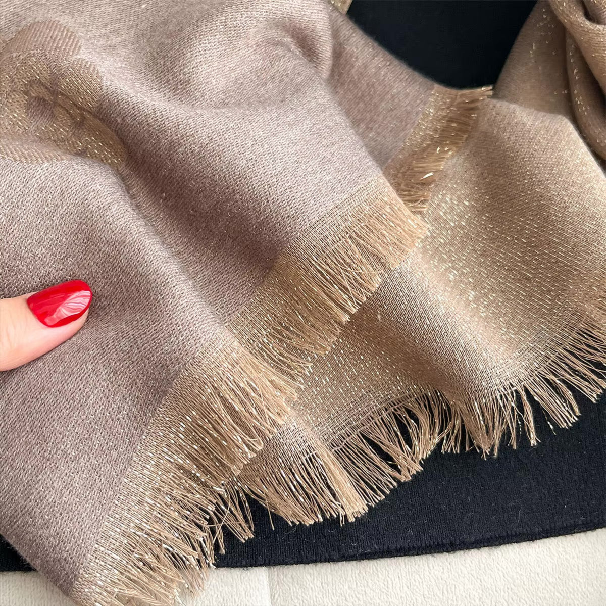 A close-up of a soft woven scarf in taupe-beige with shimmering metallic flecks and fringed edges, held by a hand with red painted nails against a dark background.