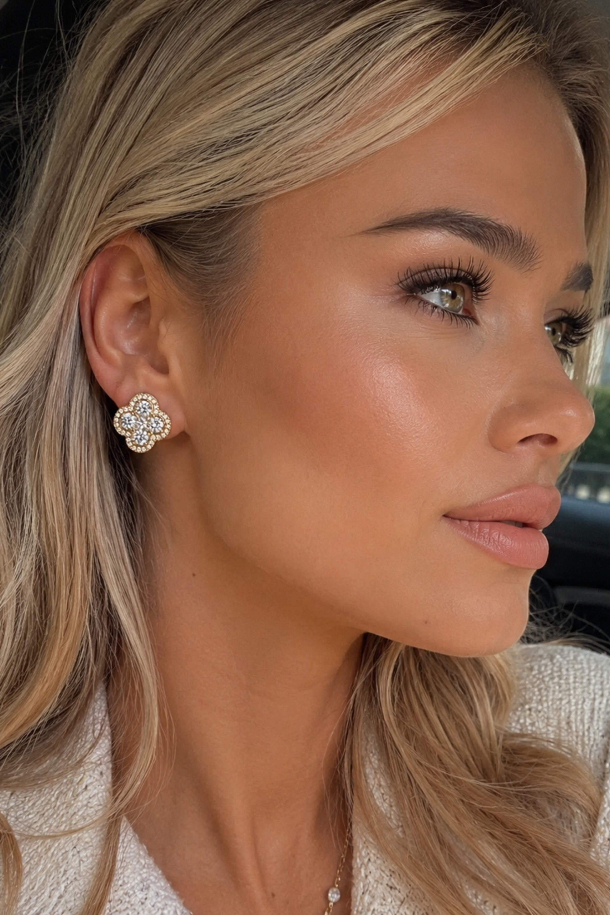 A close-up of a woman wearing a small, flower-shaped stud earring with a silver-toned metal setting and clear stones, photographed inside a car.
