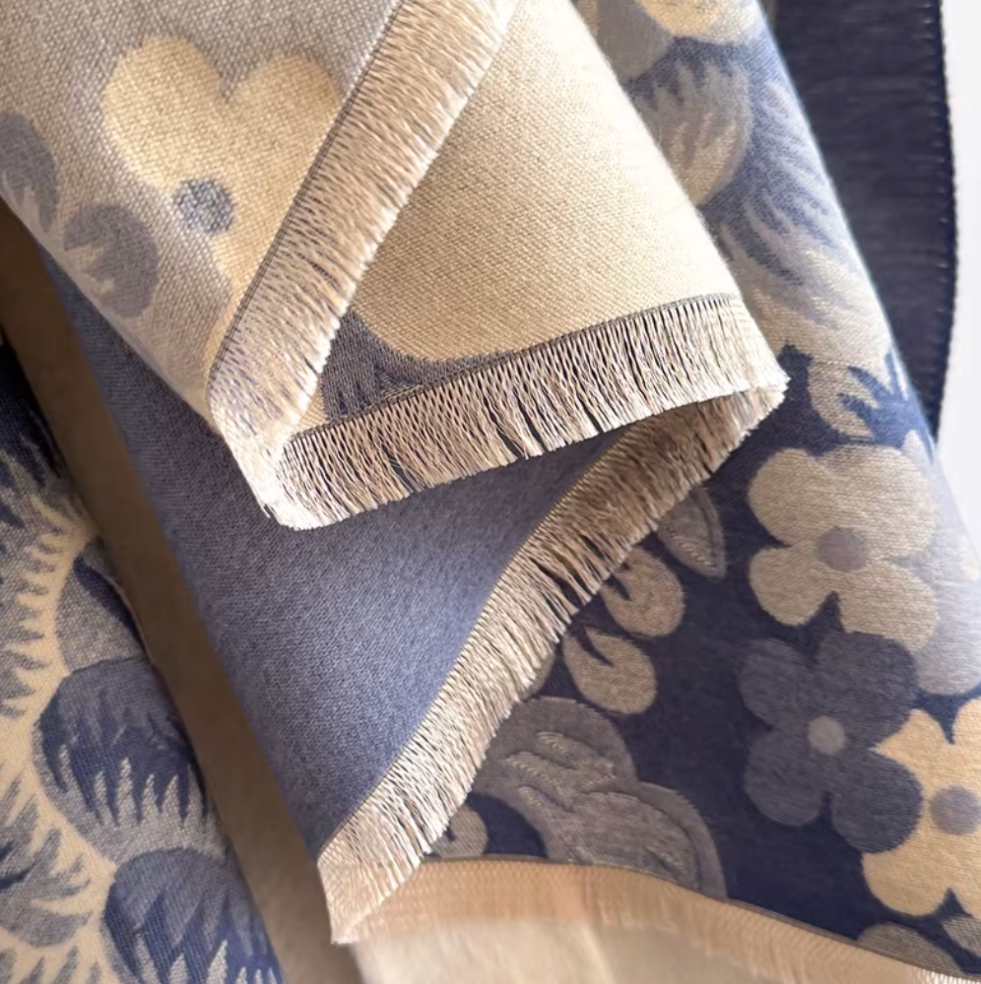 A close-up of a woven textile with a floral pattern in beige and slate blue, showing folded edges and fringed tassels.