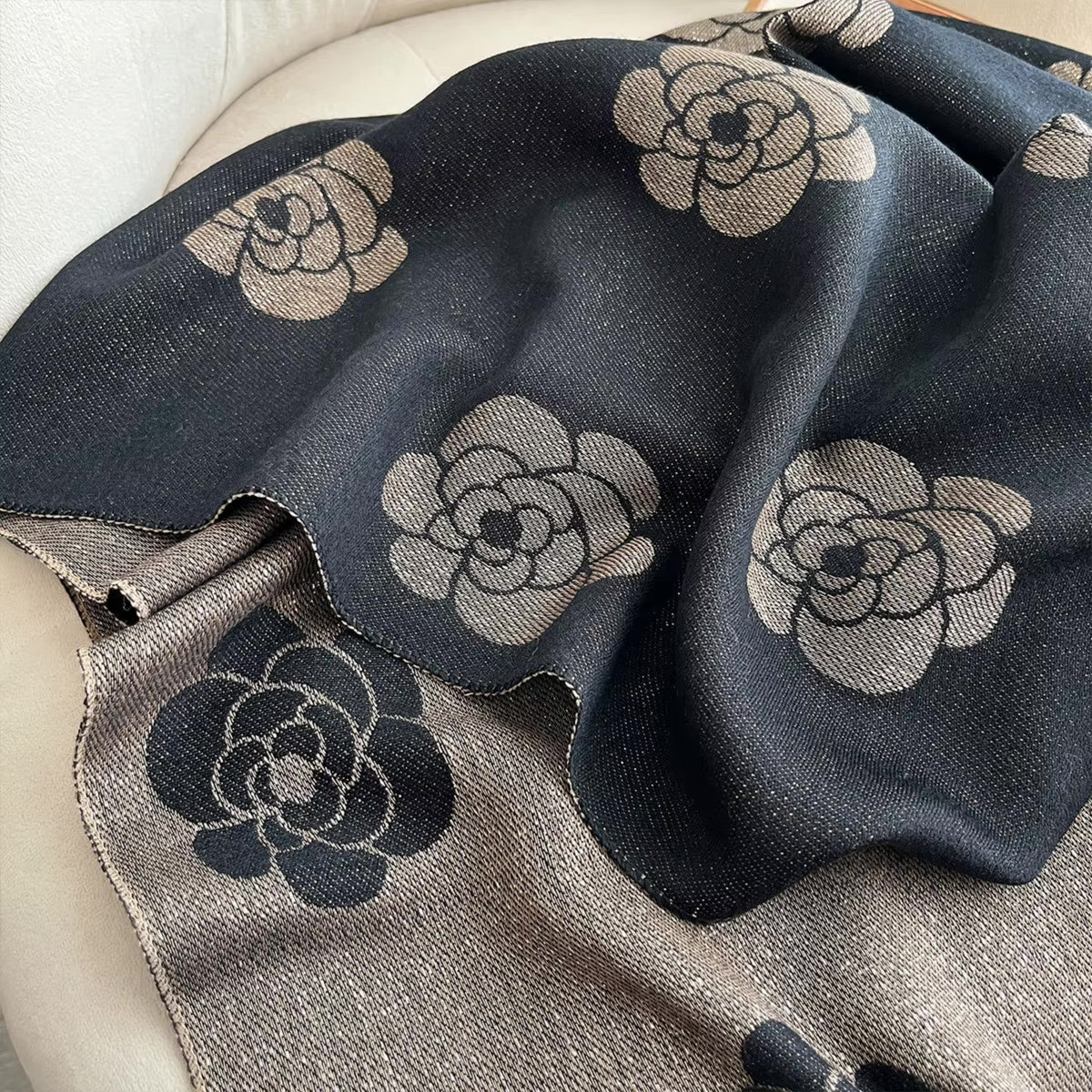A close-up of a woven throw blanket draped over a light upholstered chair. The blanket has a dark charcoal base with large beige and taupe floral motifs, and a reverse side in taupe with dark gray floral patterning.