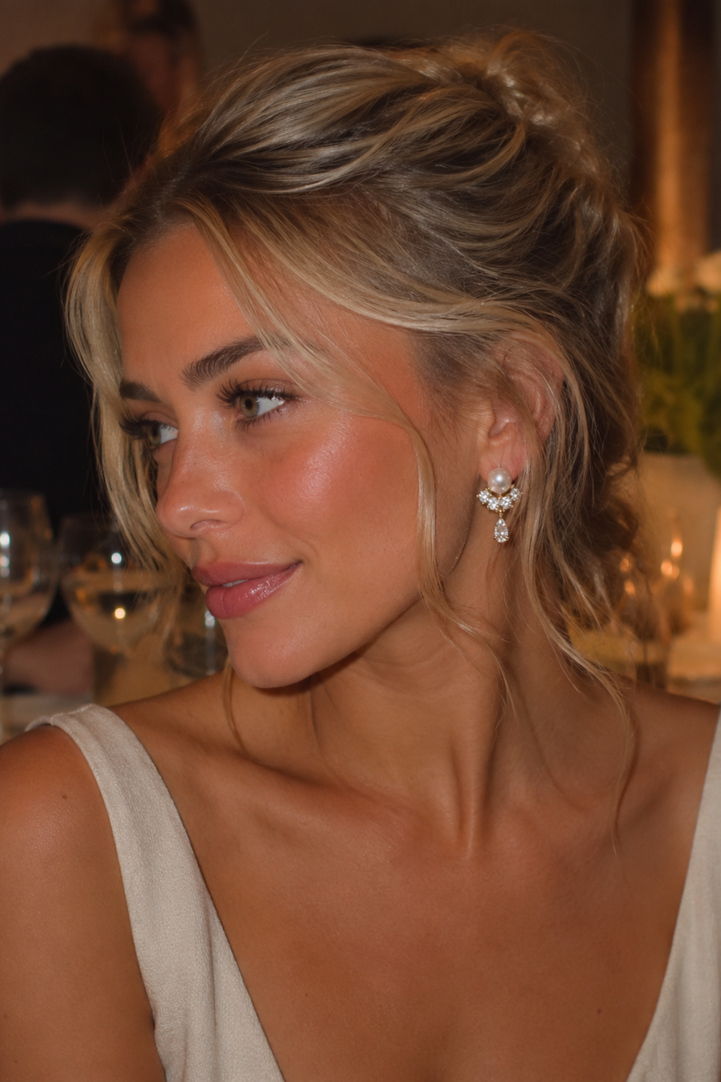 A close-up portrait of a woman with blonde hair in a half-up bun, wearing a white sleeveless top and a sparkling diamond-like drop earring in a warmly lit indoor setting.